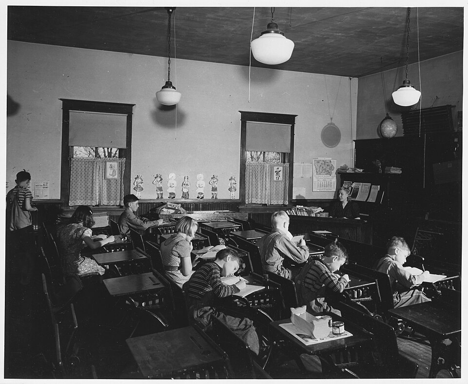 A 19th-century one-room schoolhouse with a single teacher's desk facing rows of benches.